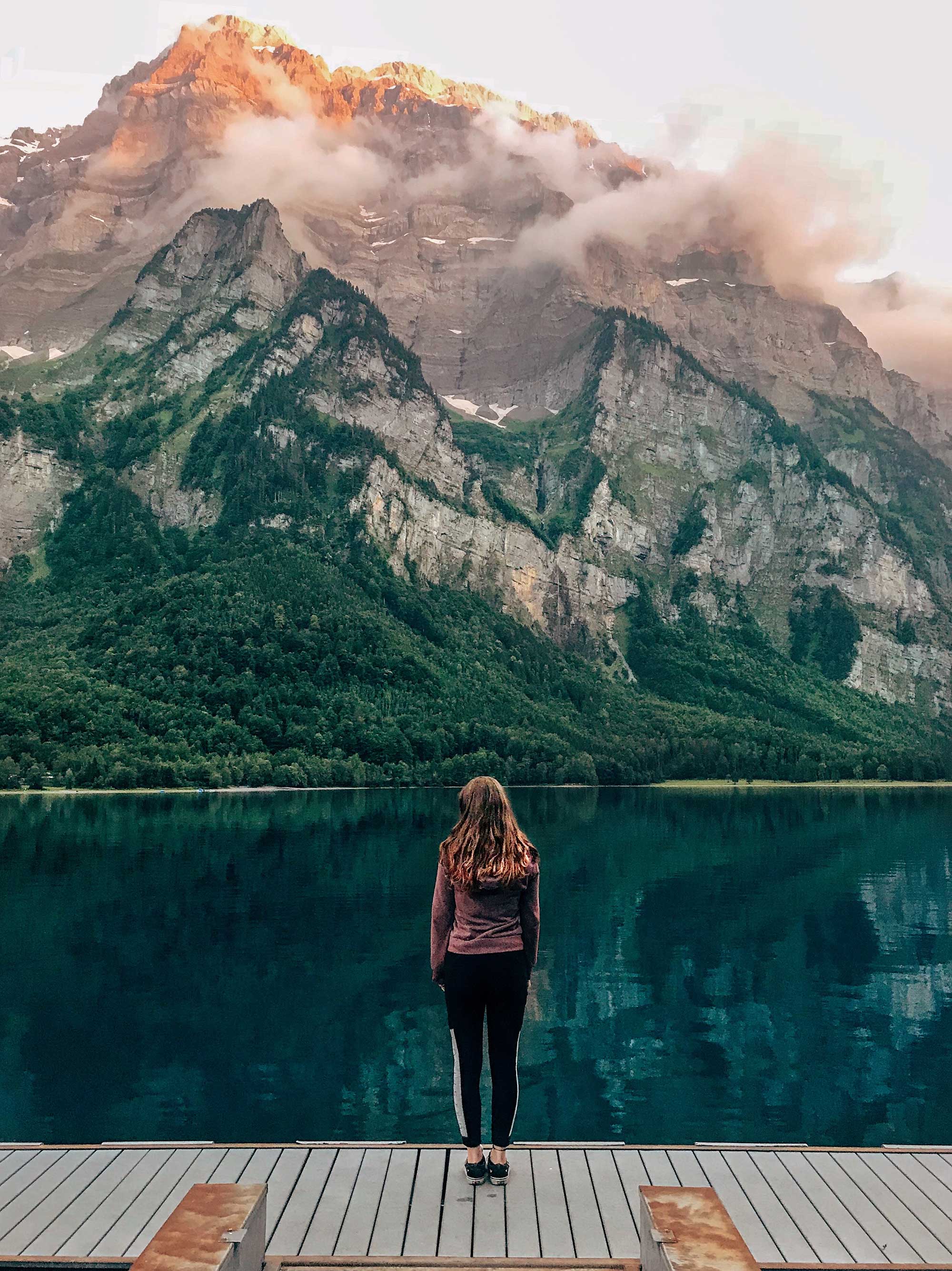 A person standing on a dock looking at a mountain.
