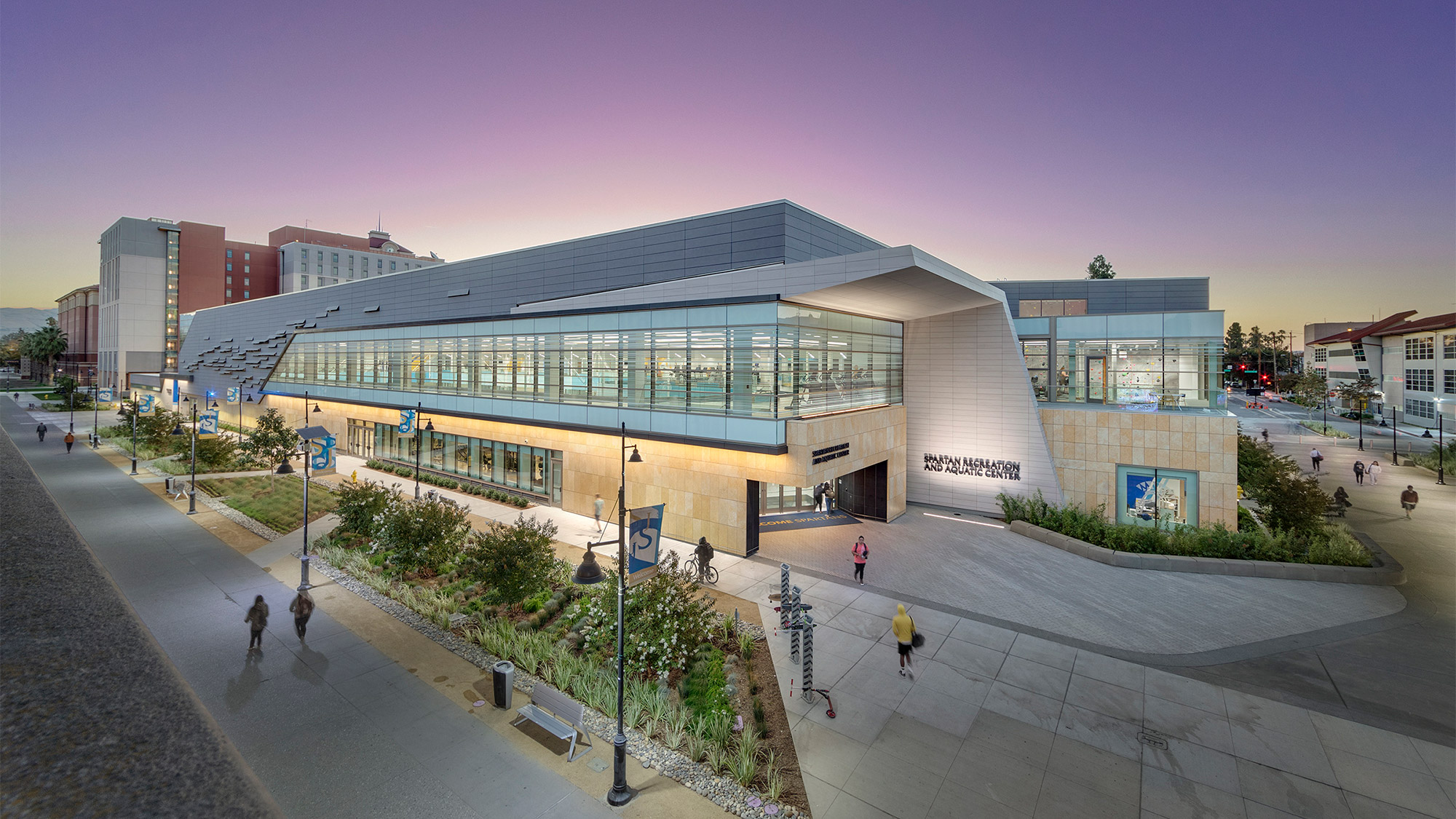 An exterior photo of a university aquatics and recreation center at dusk, showing people walking and biking to the rest of campus