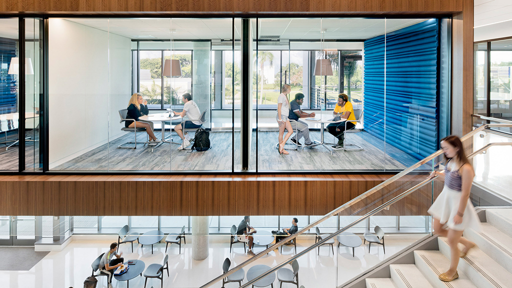 A person walking in a room with people sitting at tables and chairs.